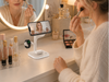 A woman follows a makeup tutorial on her phone while using an EXW 3-in-1 wireless charging station to charge her phone, smartwatch, and earbuds on an organized vanity.
