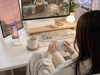 A woman relaxing in a soft chair while playing a cozy life simulation game at a cream-colored gaming desk with dual monitors, a keyboard, warm desk lighting, a PC and a white wireless charging stand.